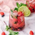 A refreshing mint mocktail, this berry mojito is garnished with lime, fresh mint, and raspberries, served in a glass on a marble surface.