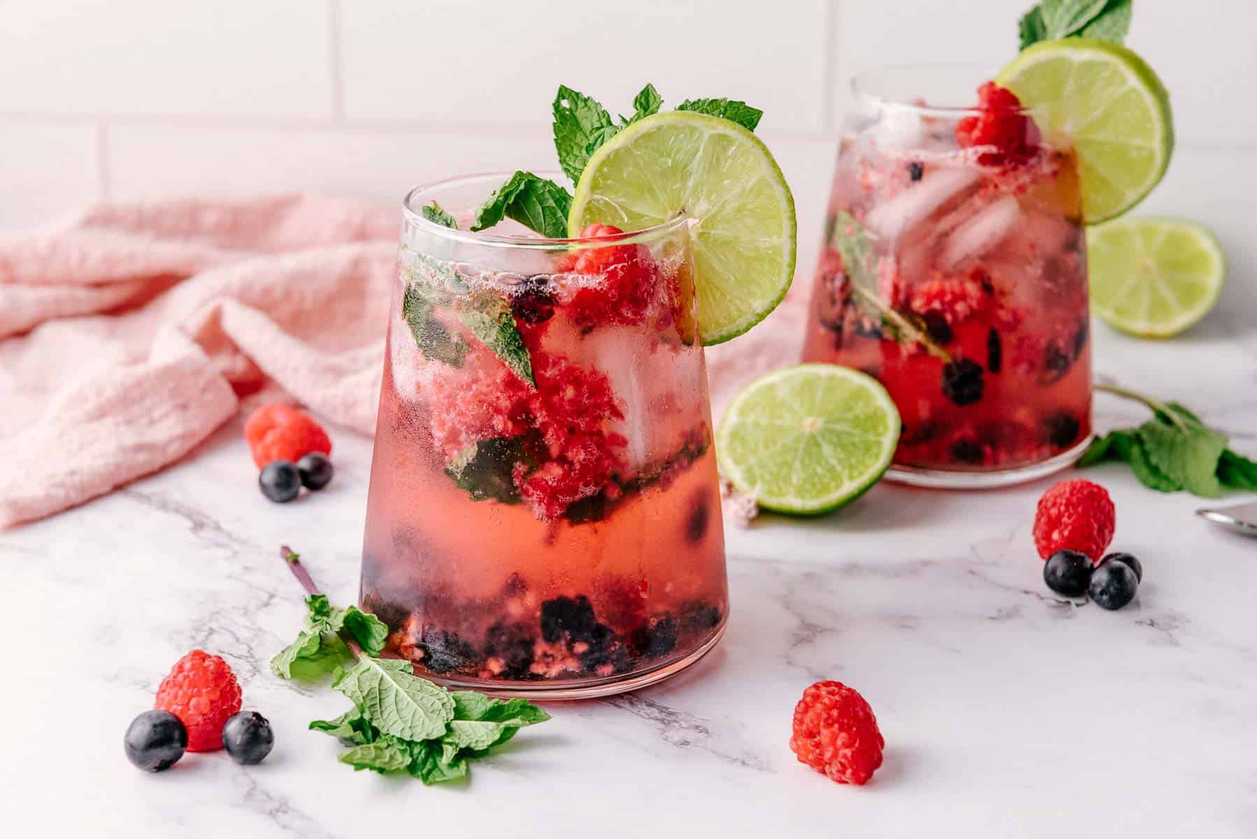 Two refreshing berry mojito cocktails with lime slices, fresh mint, blueberries, and raspberries on a marble surface&mdash;a perfect Mint Mocktail bursting with flavor.