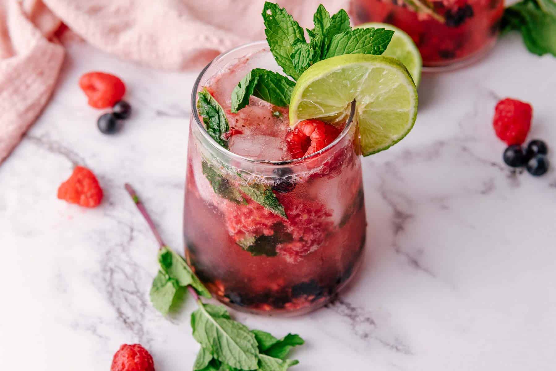 A refreshing Mint Mocktail with ice, fresh mint, lime slices, raspberry, and blueberry in a glass on a marble surface.