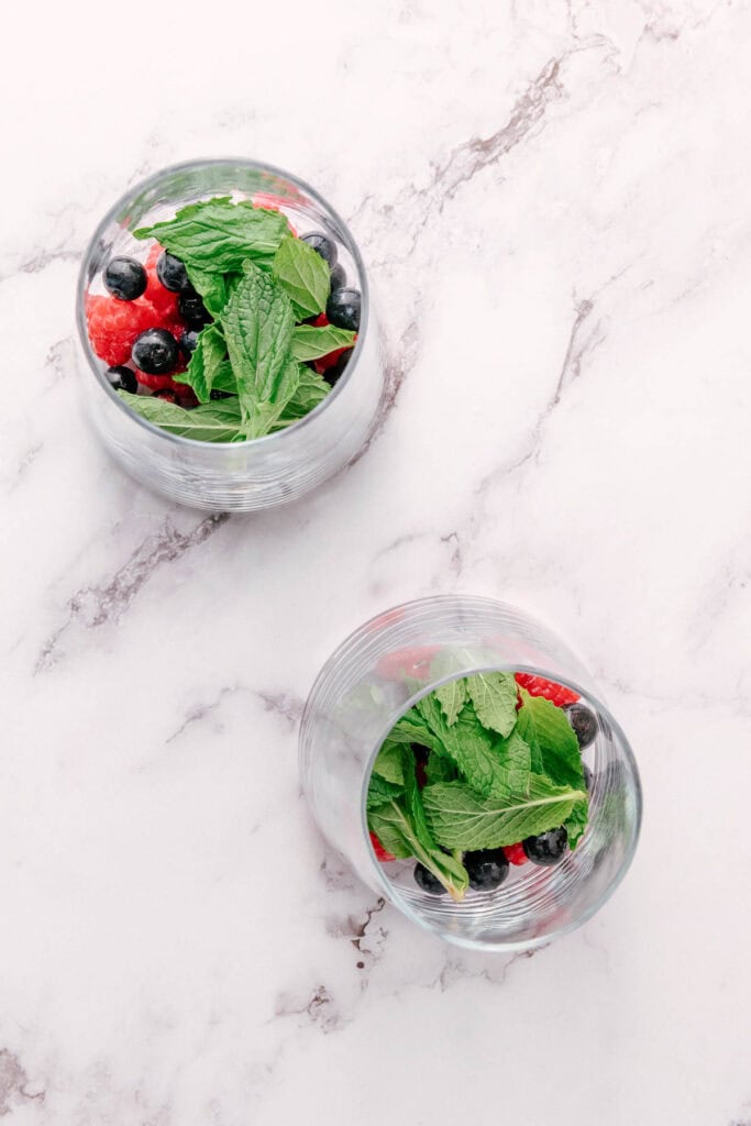 Two glasses filled with fresh mint leaves, blueberry, and raspberry create a vibrant Mint Mocktail on a white marble surface.