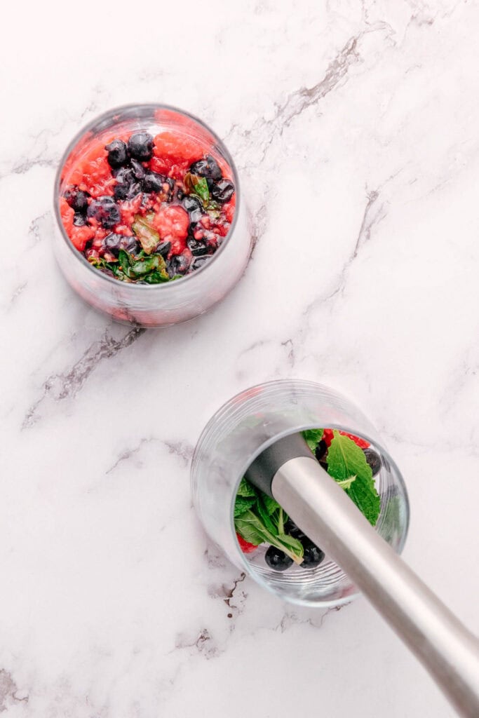 Two glasses with berries and mint, one being muddled with a metal muddler on a white marble surface&mdash;perfect for crafting a refreshing Blueberry Mint Mocktail.