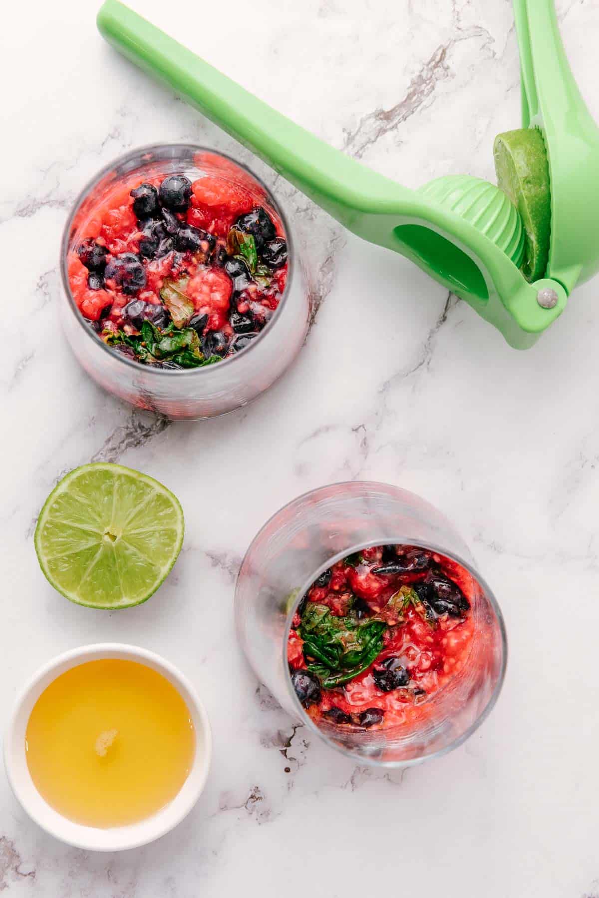 Two glasses with fruit and mint, a sliced lime, a citrus squeezer, and a bowl of honey on a marble surface&mdash;perfect for crafting a refreshing raspberry mint mocktail.