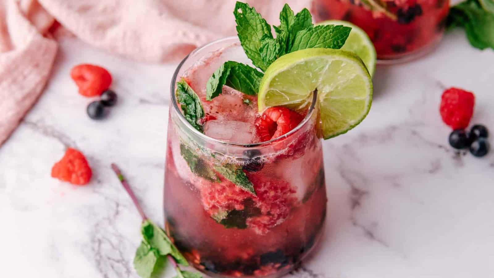 A berry mojito with lime slices, mint leaves, and ice in a glass on a marble surface.
