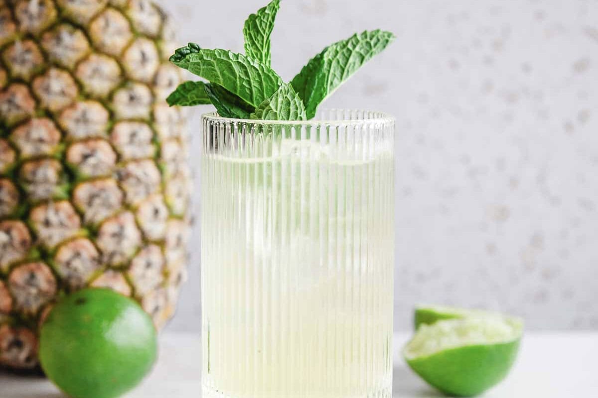 A glass of iced drink with mint leaves, next to a pineapple and a halved lime on a light surface, perfect for a refreshing Tropical Electrolyte Mocktail Recipe.