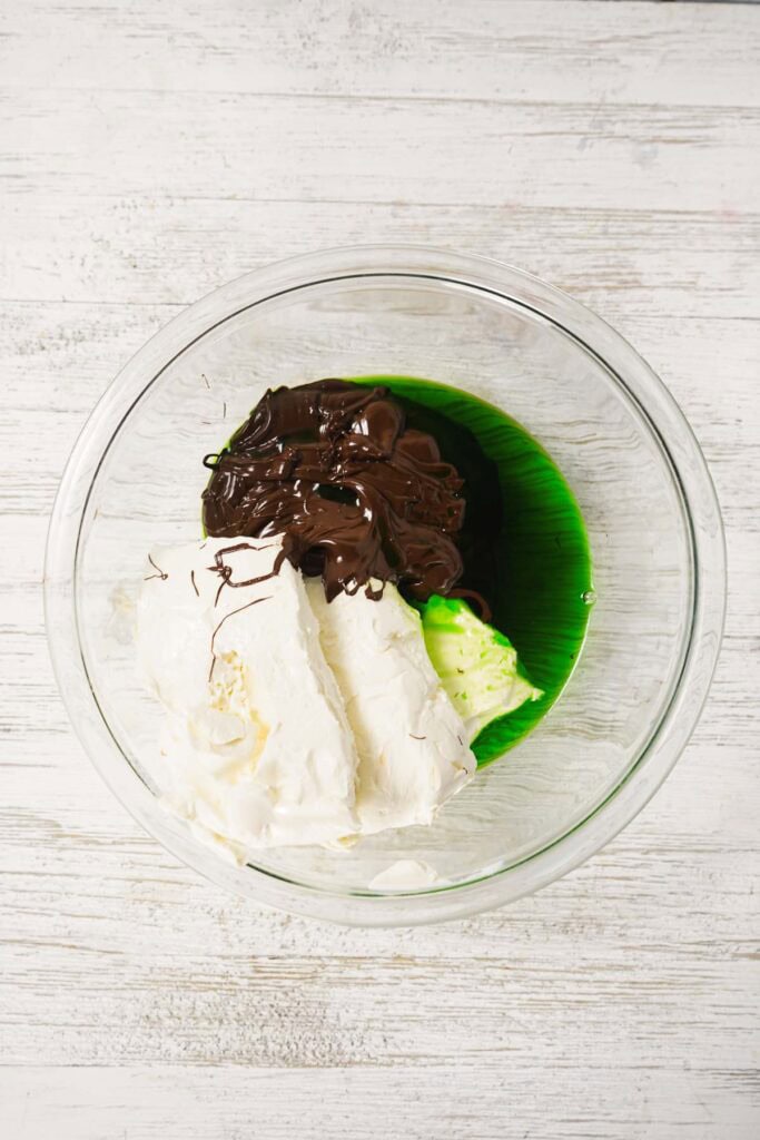 A glass bowl with mint cream cheese, green liquid, and melted chocolate on a white wooden surface&mdash;perfect for making easy dessert chocolate truffles.