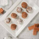Eight chocolate cream cheese truffles, some dusted with powdered sugar, rest on a white plate with wafers and a spoon of sugar nearby.