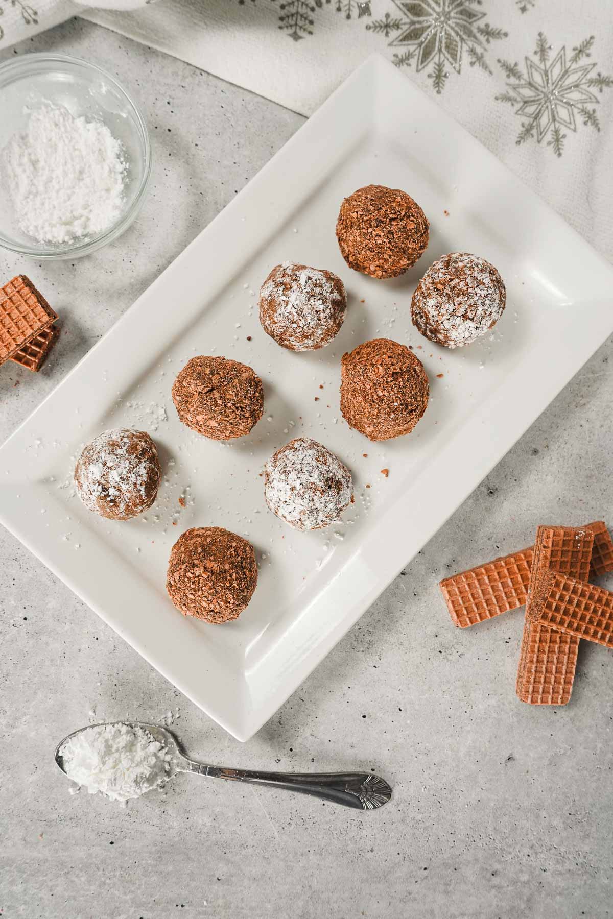 Eight chocolate cream cheese truffles, some dusted with powdered sugar, rest on a white plate with wafers and a spoon of sugar nearby.