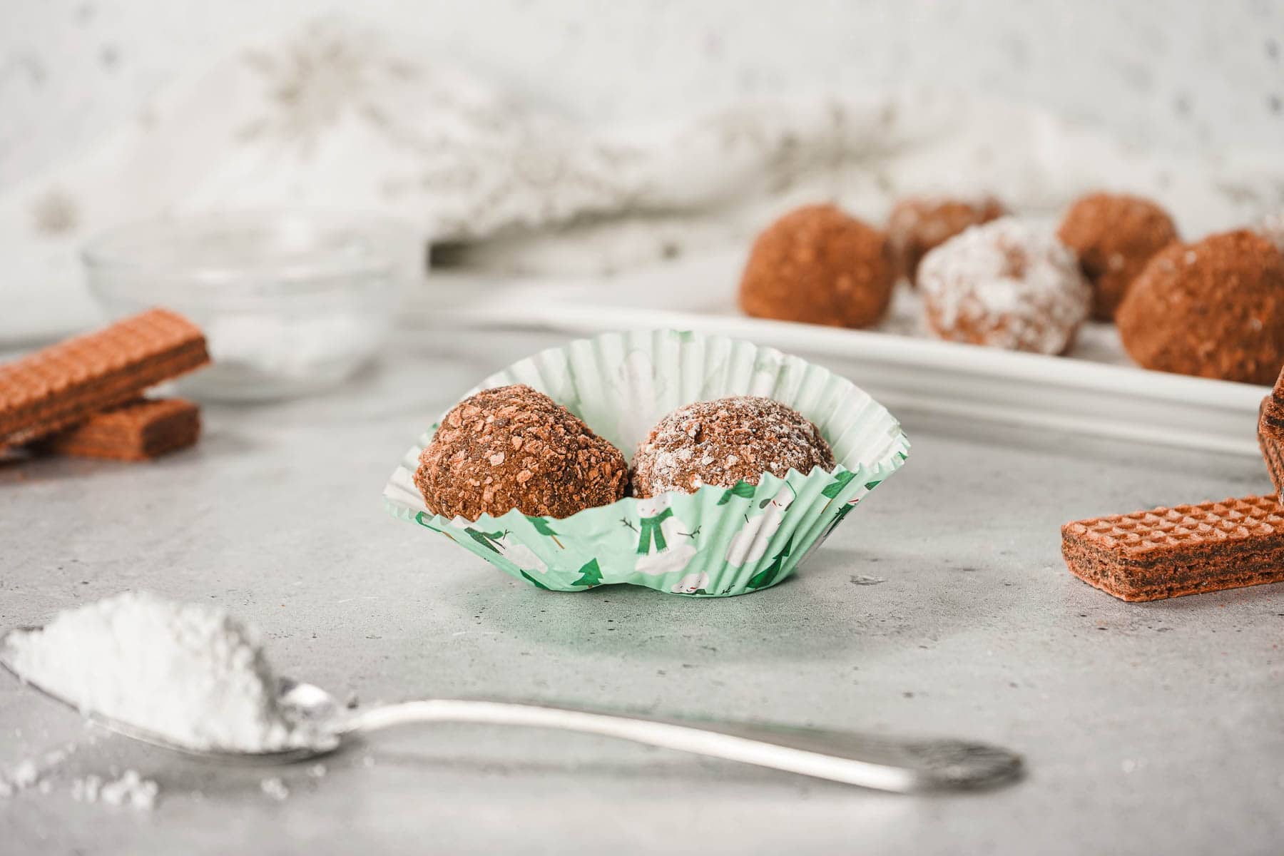 Two Cream Cheese Truffles in a paper cup, with wafers and powdered sugar on a gray surface&mdash;an easy truffle recipe perfect for any occasion.