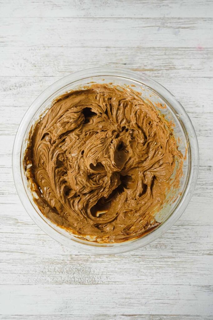 A glass bowl filled with swirled chocolate frosting on a white wooden surface, perfect for topping Chocolate Mint Truffles or as a base for your favorite Easy Truffle Recipe.