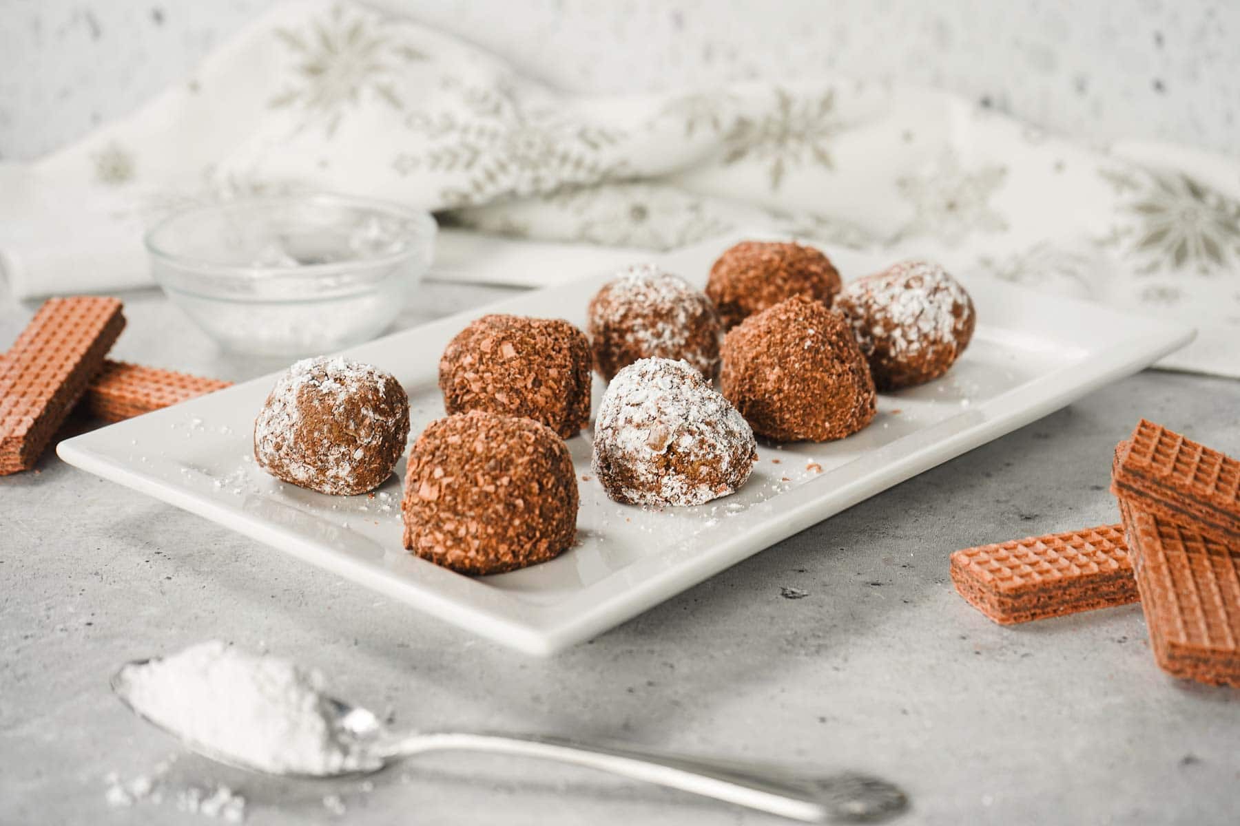Chocolate truffles with powdered sugar on a white plate, surrounded by wafer cookies and a spoon of sugar&mdash;an easy truffle recipe that's perfect for any occasion.
