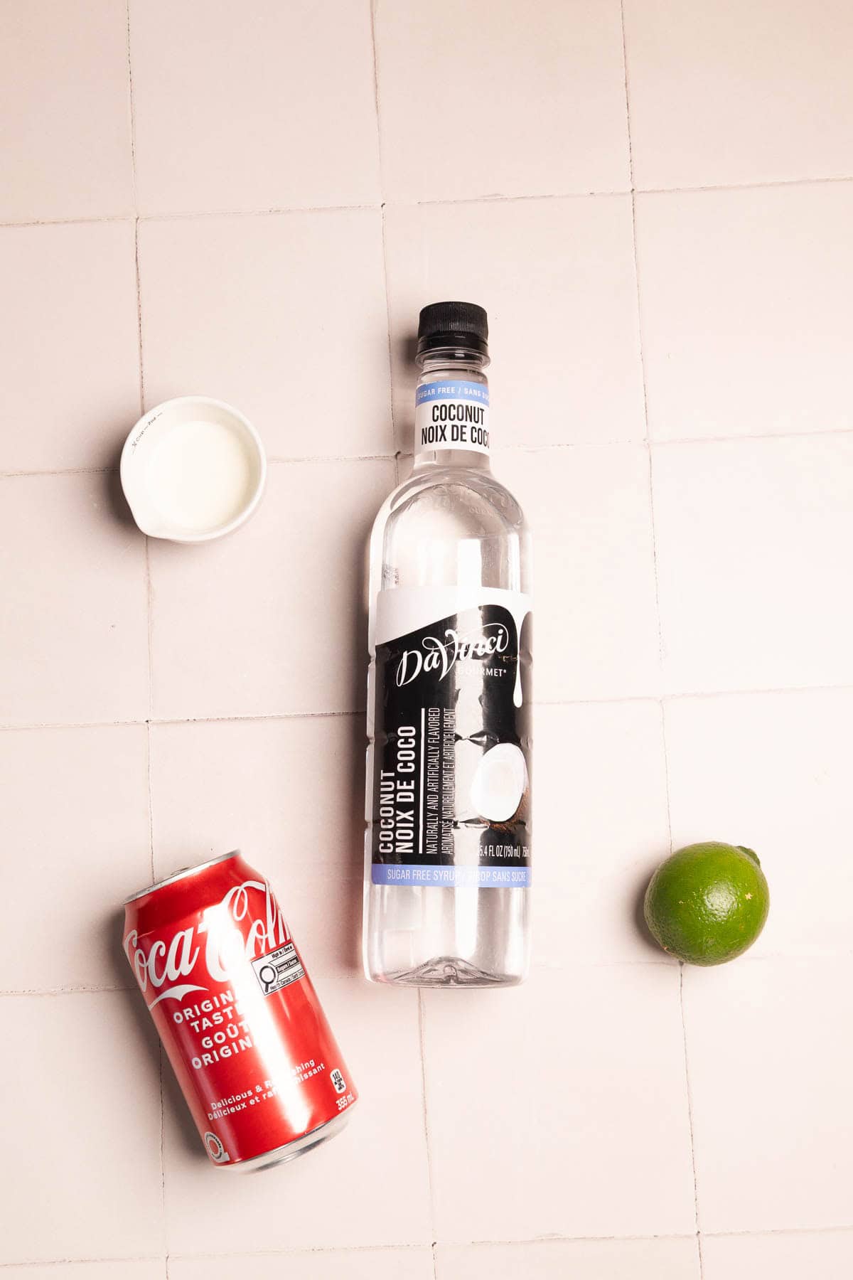 A can of Coca-Cola, coconut vodka, a lime, and a bottle cap on a tiled surface&mdash;perfect ingredients for a refreshing Dirty Soda or creative mocktail recipe with coconut flair.