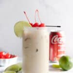 A creamy coconut iced drink in a glass with cherries and lime, accompanied by a Coke can, limes, and cherries—perfect for trying as a refreshing Dirty Soda mocktail recipe.