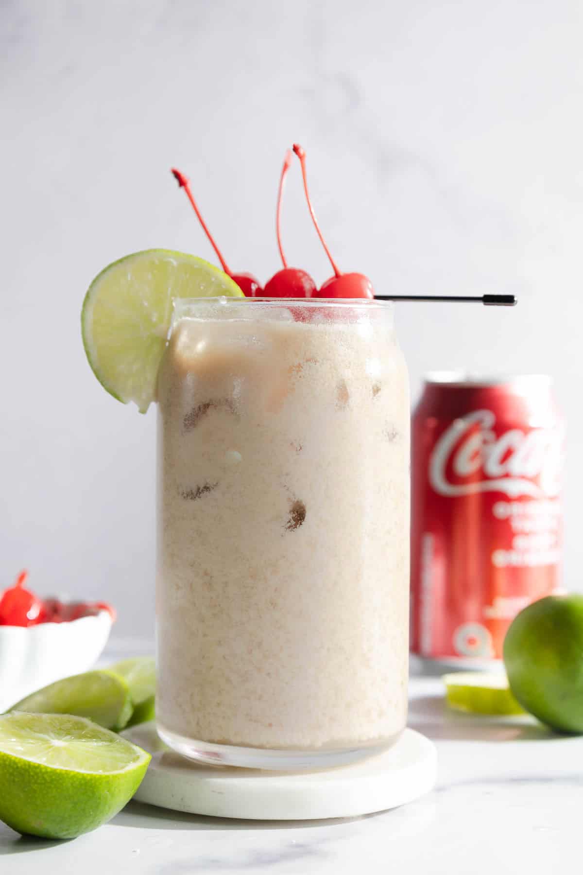 A frosty Dirty Soda mocktail with ice, cherries, a lime slice garnish, and a hint of coconut, served alongside a can of Coca-Cola and fresh limes.