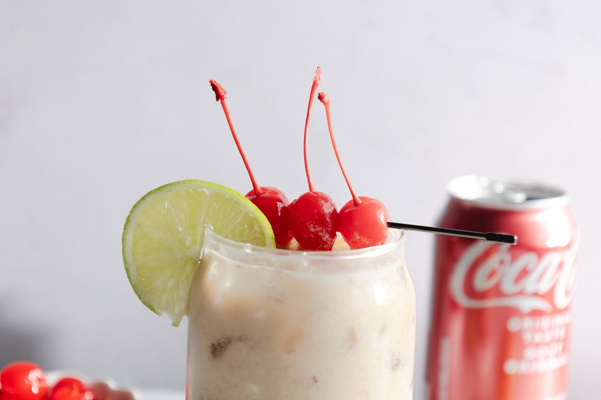A creamy coconut mocktail recipe served over ice, topped with cherries and a lime slice, with a Coca-Cola can adding Dirty Soda vibes in the background.