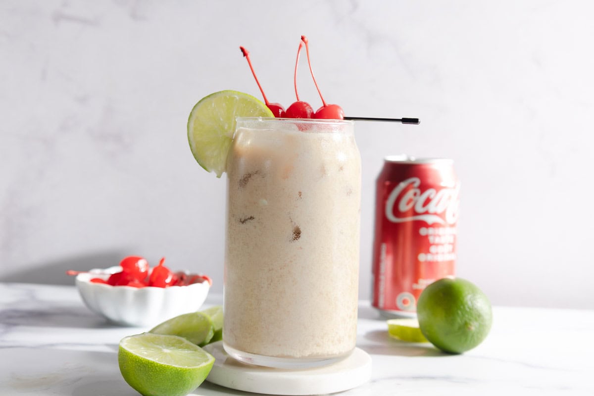 A creamy coconut drink with ice, lime wedge, and cherries in a glass, accompanied by a Coca-Cola can and limes&mdash;perfect for trying the Dirty Soda mocktail recipe.
