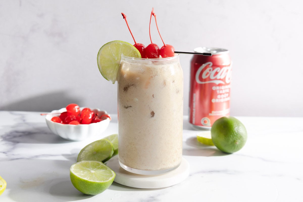 A creamy coconut iced drink, inspired by the Dirty Soda mocktail recipe, garnished with cherries and a lime slice, with a can of Coca-Cola and fresh limes nearby.