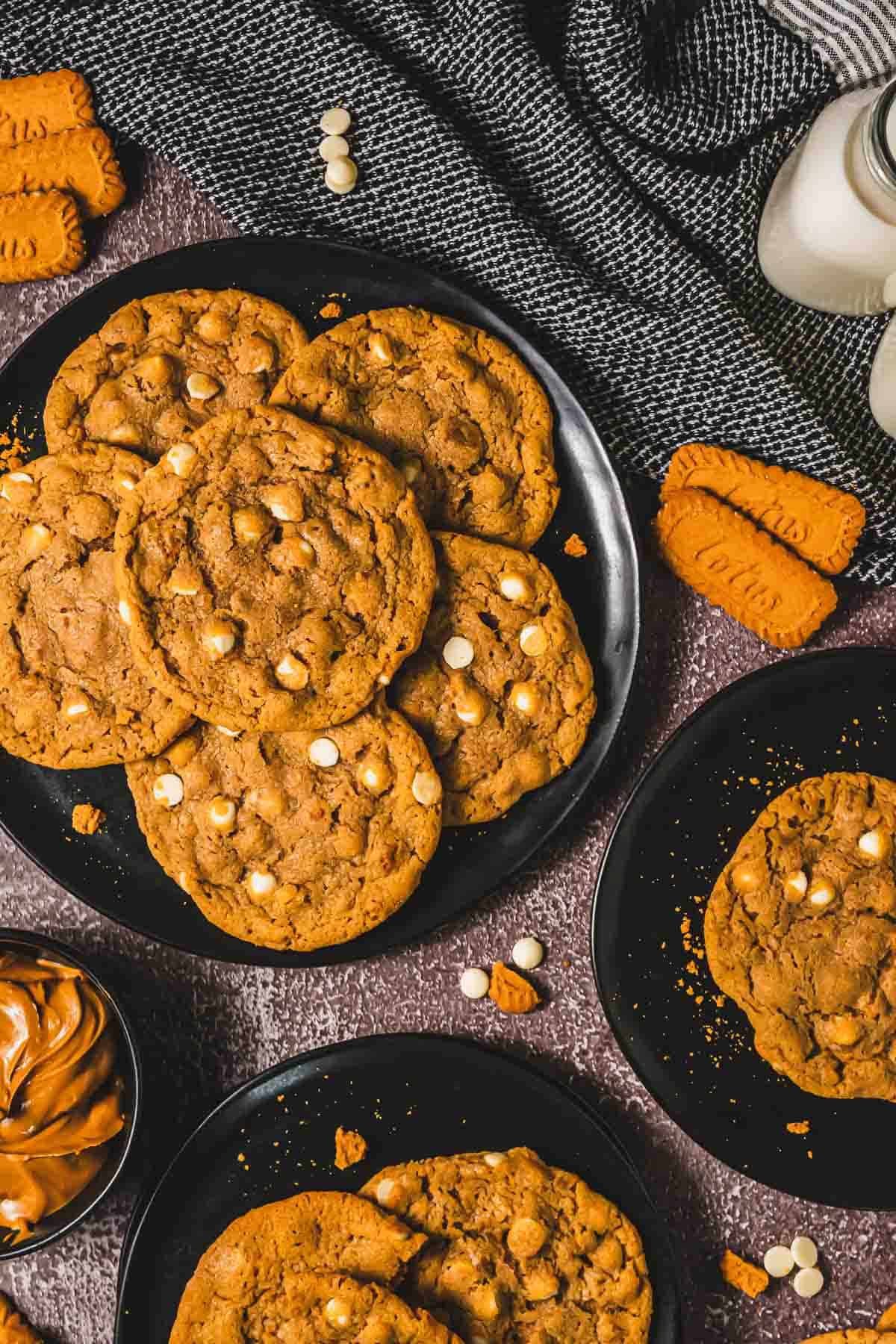 A plate of cookies with white chocolate chips, inspired by Crumbl Biscoff, surrounded by Biscoff cookies, milk, and a bowl of spread.