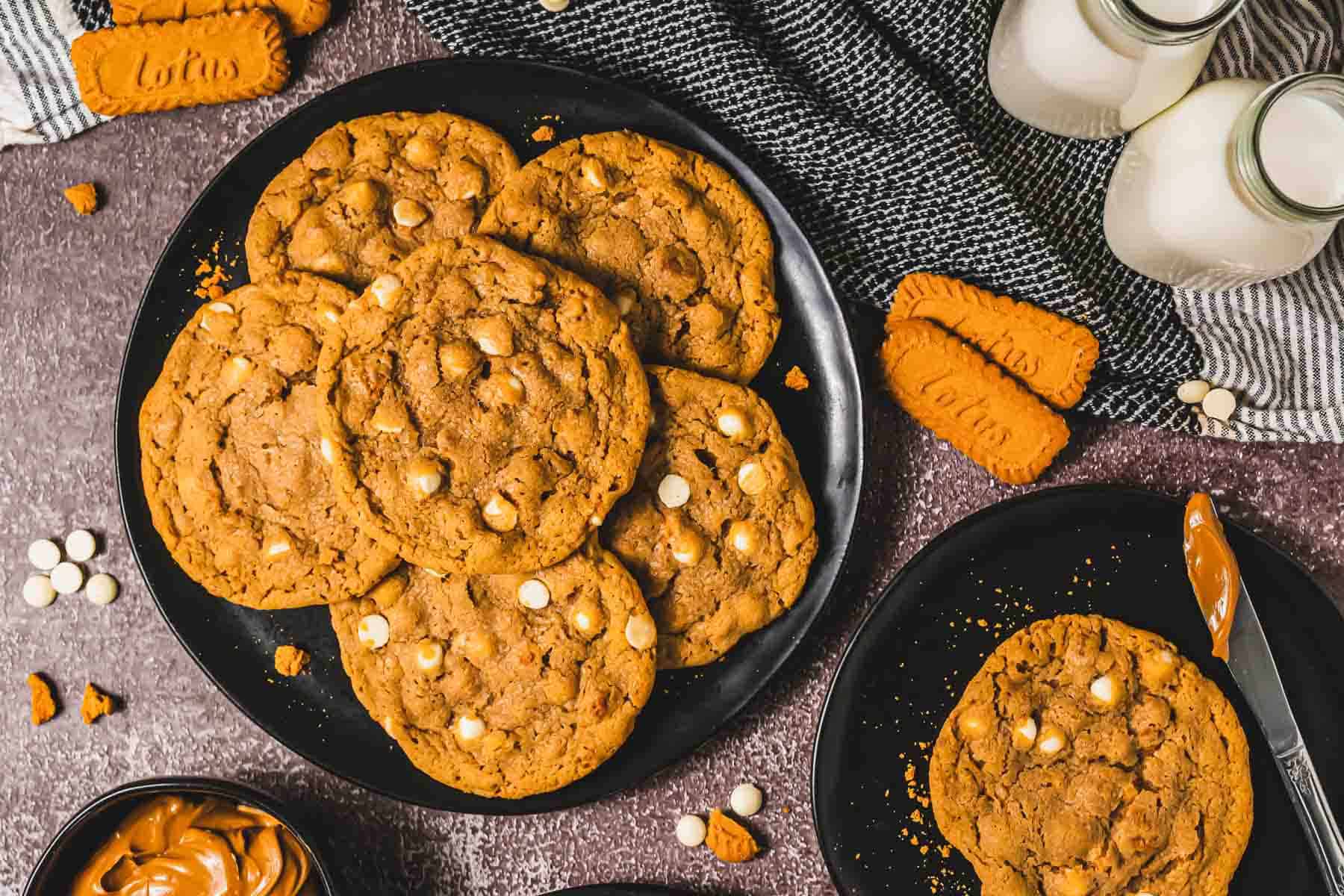 A plate of Copycat Biscoff Cookies with white chocolate chips, surrounded by Lotus cookies, milk bottles, and a bowl of spread.