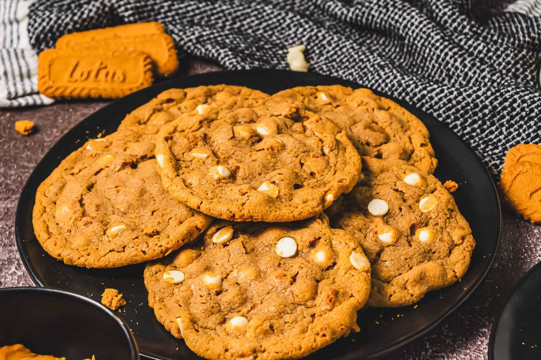 A plate of cookies with white chocolate chips, surrounded by Biscoff cookies, sits on a textured surface&mdash;perfect for fans of Crumbl Biscoff or anyone craving a delicious Crumbl copycat cookie experience.