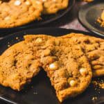 A large Crumbl Copycat cookie with white chocolate chips broken in half on a black plate, with more cookies in the background.