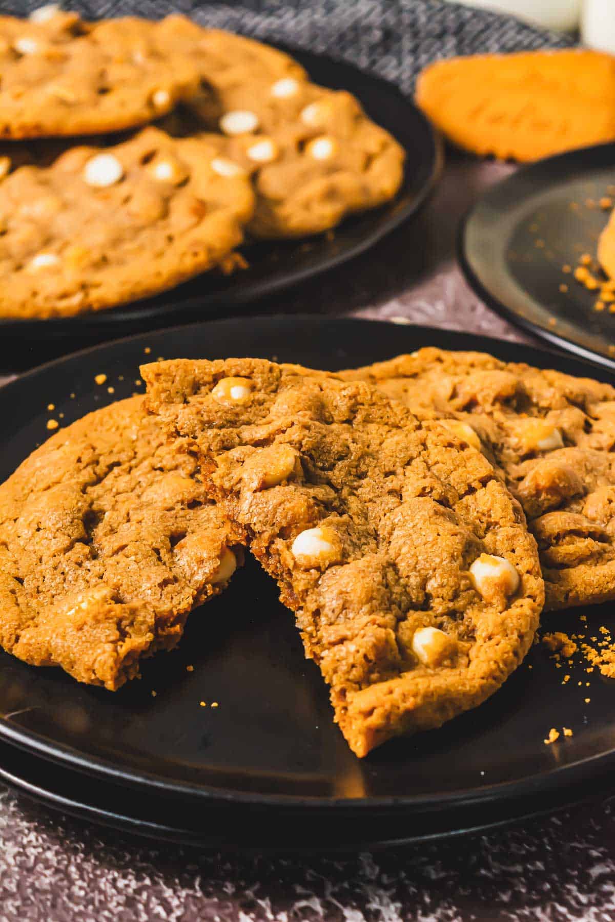 A large Crumbl Copycat cookie with white chocolate chips broken in half on a black plate, with more cookies in the background.