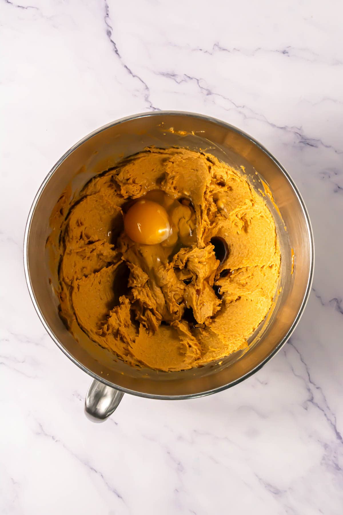 A metal mixing bowl with cookie dough and a cracked egg on top, perfect for making delicious Crumbl copycat treats, sits on a marble countertop.