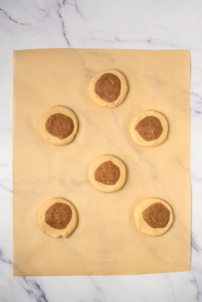 Six round cinnamon swirl cookies with dark centers rest on parchment paper, arranged neatly on a light marble surface.