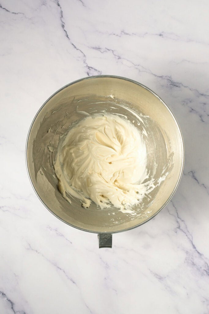 A metal mixing bowl with creamy white frosting on a marble surface, perfect for topping homemade Crumbl copycat cookies.