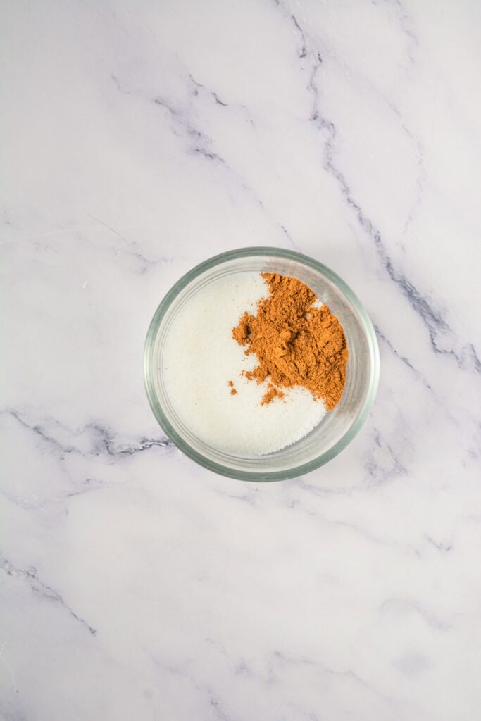 A glass bowl with white sugar and brown cinnamon on a marble surface&mdash;perfect for making cinnamon swirl cookies or your favorite Crumbl copycat treats.