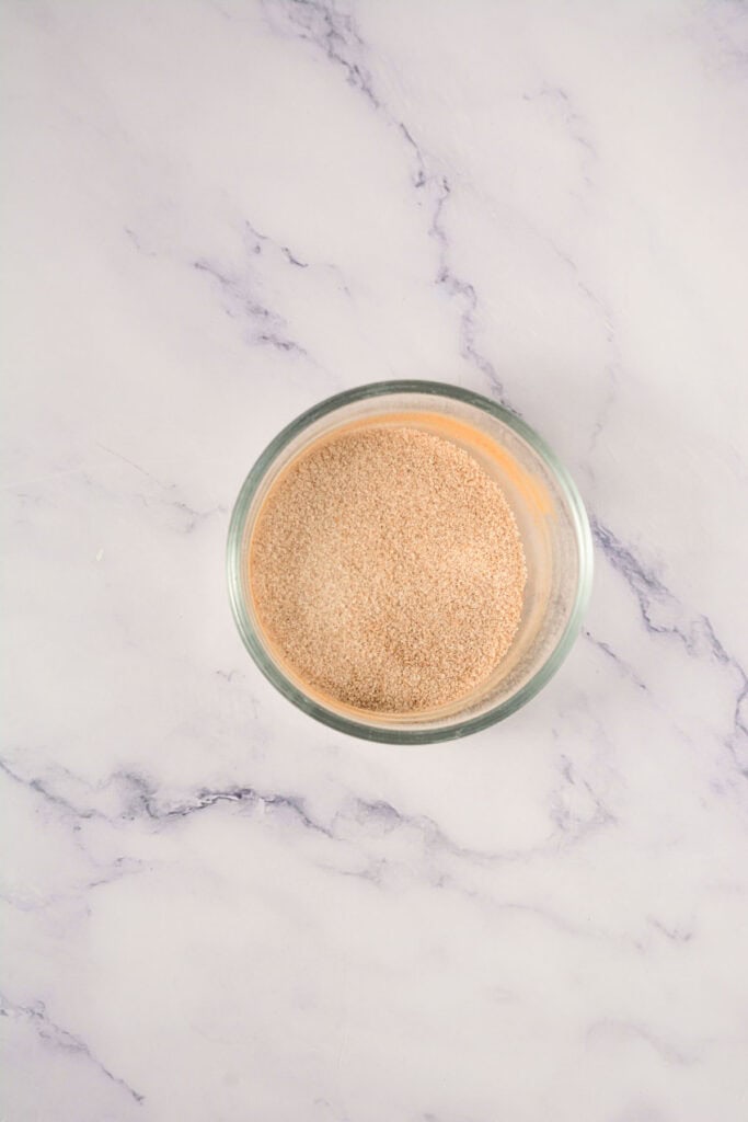 A glass bowl filled with dry yeast sits on a light marble surface, ready to help you create delicious Crumbl Copycat cinnamon swirl cookies at home.