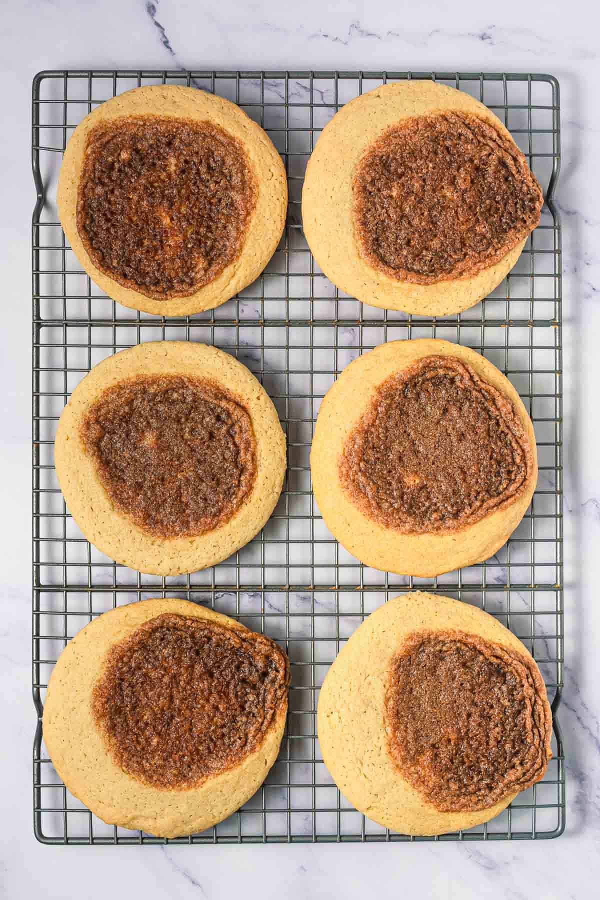 Six large cinnamon swirl cookies with brown sugar cinnamon centers cooling on a wire rack over a marble surface&mdash;perfect Crumbl copycat cookies for any sweet craving.