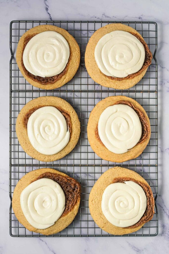 Six large cinnamon swirl cookies with white icing on a cooling rack over a marble surface&mdash;perfect Crumbl copycat cookies for any sweet tooth.