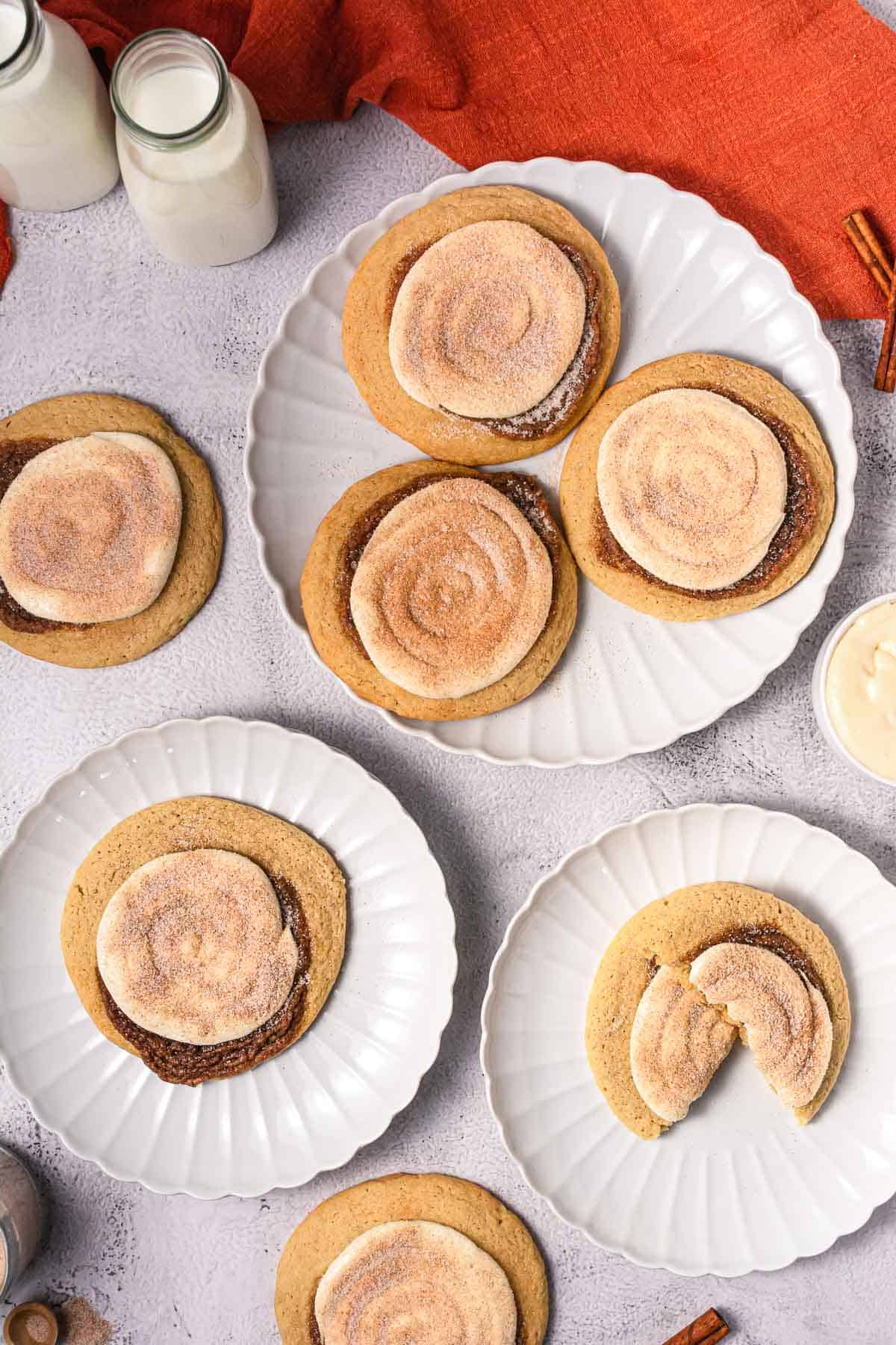 Five plates with large cinnamon swirl cookies topped with cinnamon sugar, next to bottles of milk and an orange napkin&mdash;perfect for anyone craving a Crumbl copycat cookie recipe.