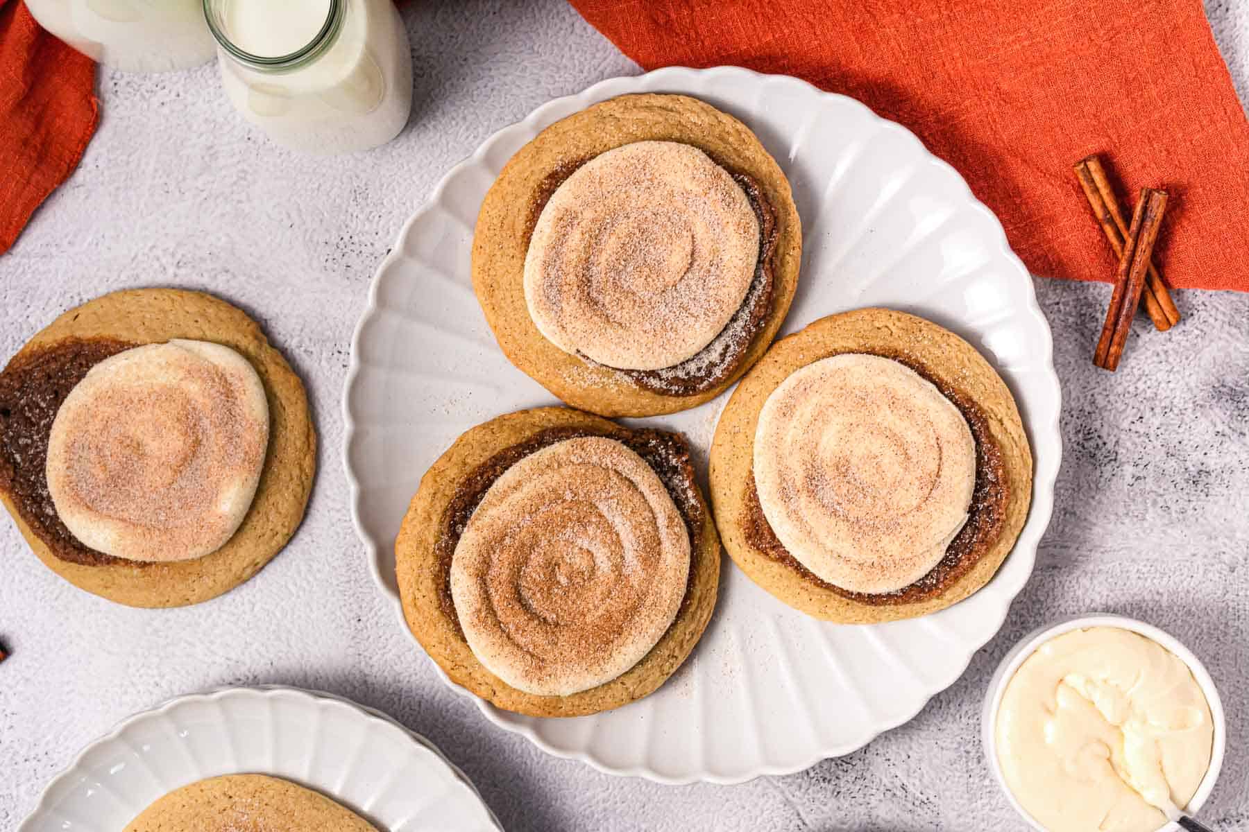 Four Crumbl copycat cinnamon roll cookies with icing and cinnamon sugar on a white plate, surrounded by milk and cinnamon sticks.