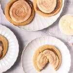Cinnamon swirl cookies topped with cinnamon sugar and frosting are arranged on white plates, with one Crumbl copycat cookie broken in half.