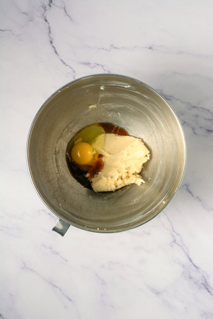 A metal mixing bowl with sugar, butter, an egg, and vanilla extract on a marble countertop&mdash;perfect for whipping up Crumbl copycat cinnamon swirl cookies.