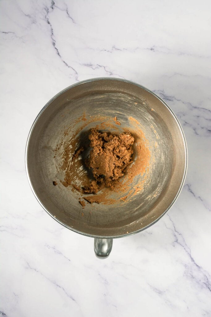 A metal mixing bowl with brown cookie dough, perfect for making cinnamon swirl cookies, sits on a white marble surface.