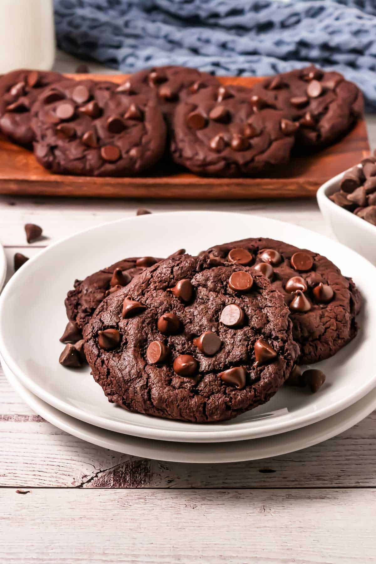 Two Copycat Cookies on a white plate, with more cookies and chocolate chips in the background&mdash;a delicious Crumbl Copycat treat.