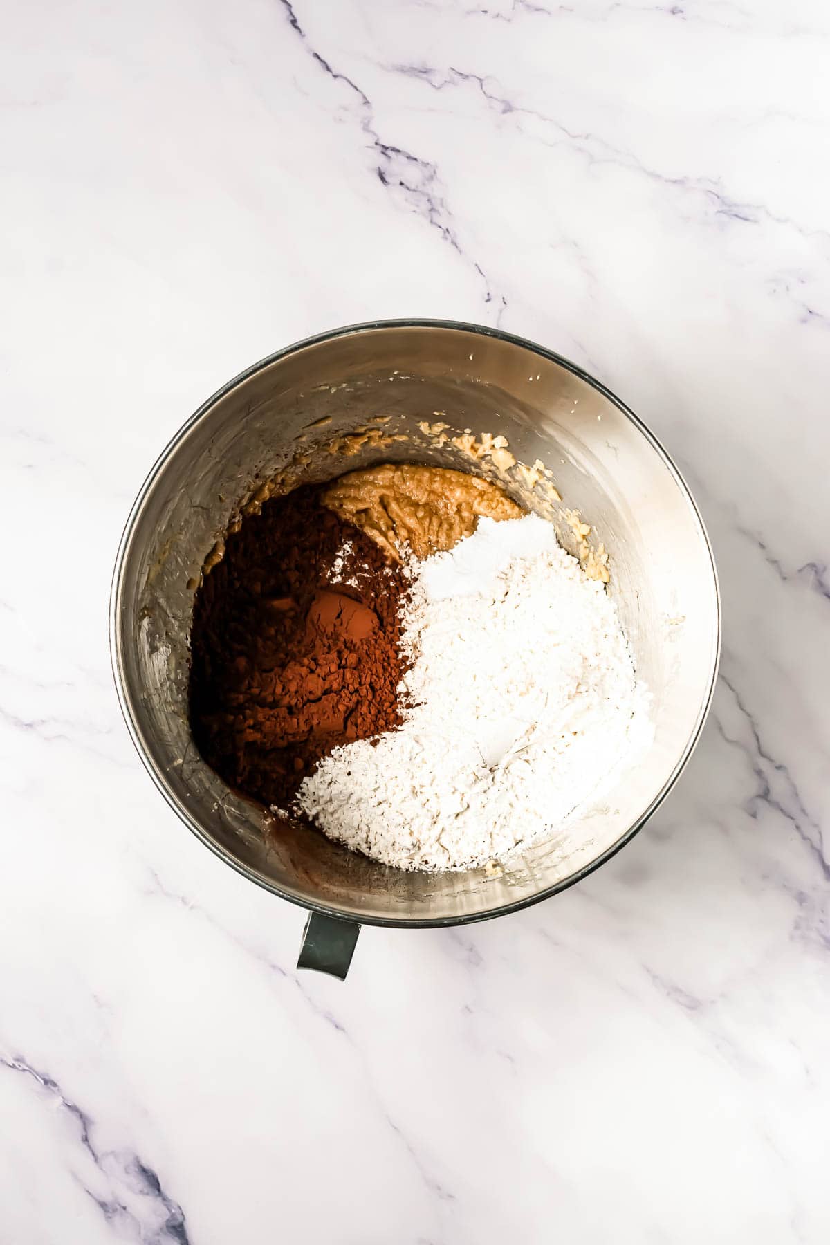 A metal mixing bowl with flour, cocoa powder, and wet ingredients on a marble surface&mdash;perfect for preparing Crumbl Copycat Dark Dream Cookies.