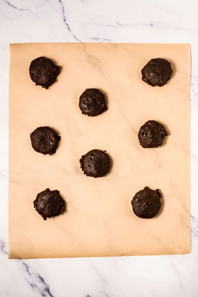 Eight scoops of chocolate cookie dough, inspired by Crumbl Copycat recipes, rest on parchment paper in three neat rows atop a marble surface.