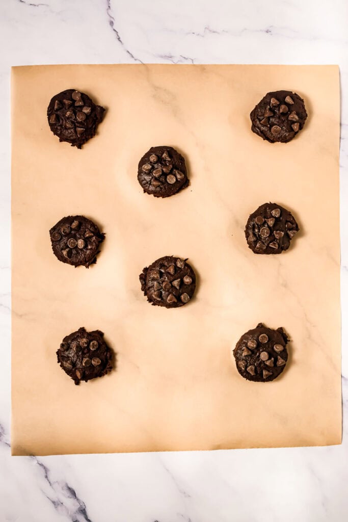 Eight unbaked Crumbl Copycat Cookies, rich with chocolate chips, are spaced on parchment paper atop a marble surface.