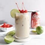 Iced creamy drink in a glass with cherries and lime, a Coke can, limes, and a bowl of cherries nearby&mdash;perfect for trying your own Coconut Dirty Soda mocktail recipe.