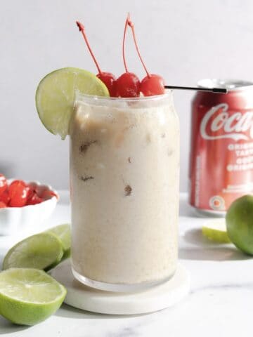 Iced creamy drink in a glass with cherries and lime, a Coke can, limes, and a bowl of cherries nearby—perfect for trying your own Coconut Dirty Soda mocktail recipe.