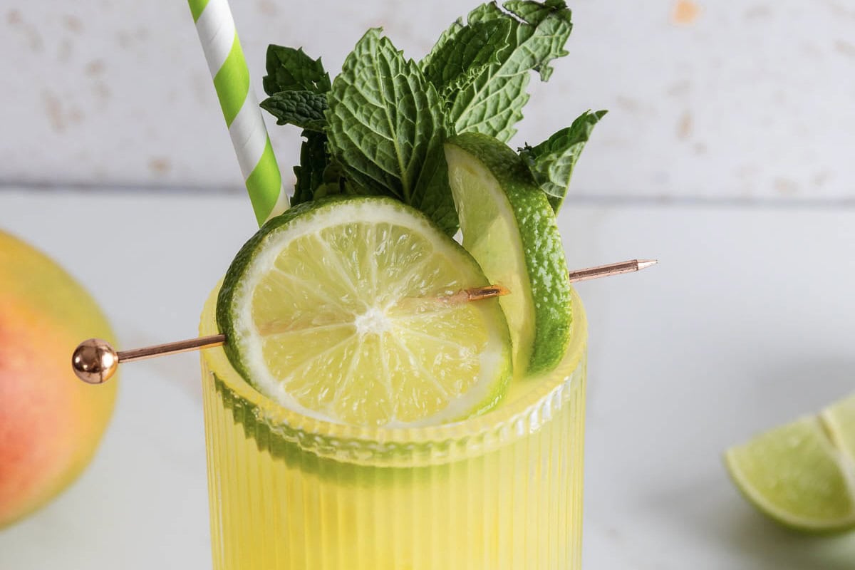 A yellow mango drink garnished with lime slices, mint leaves, and a striped straw in a glass&mdash;perfect as an electrolyte-rich mocktail recipe for any occasion.