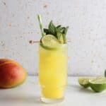 Yellow mango drink in a tall glass with lime, mint, and a striped straw; vibrant mocktail recipe features mango and lime beside the glass for a refreshing burst of flavor.