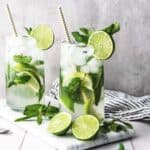 Two glasses of Faux-jito, a refreshing non-alcoholic drink with lime slices, mint leaves, ice, and straws on a marble board surrounded by limes and mint.