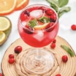 A refreshing cranberry spritzer mocktail with ice, cranberries, and mint sits on a woven mat, surrounded by citrus slices for a vibrant, non-alcoholic treat.
