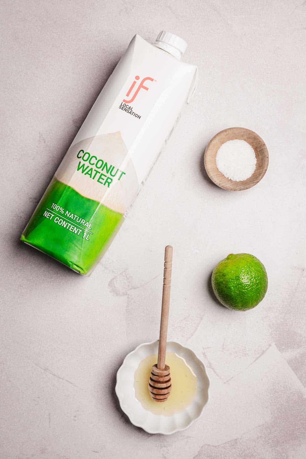 Carton of coconut water, a bowl of salt, a lime, and a dish of honey with a honey dipper on a light surface&mdash;perfect ingredients for an easy homemade electrolyte drink or refreshing mocktail recipe.