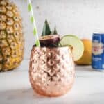 A copper pineapple cup with a green-striped straw, lime garnish, and tropical fruit sits on a white surface&mdash;perfect for serving your favorite Tropical Mocktail.