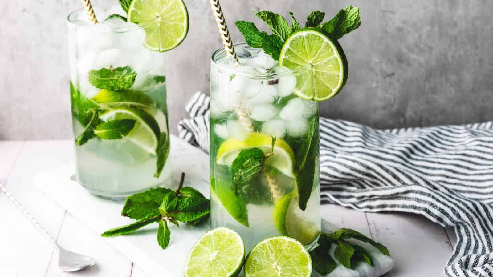 A glass of mojito with lime, mint leaves, ice, and a straw on a table with a striped cloth nearby.